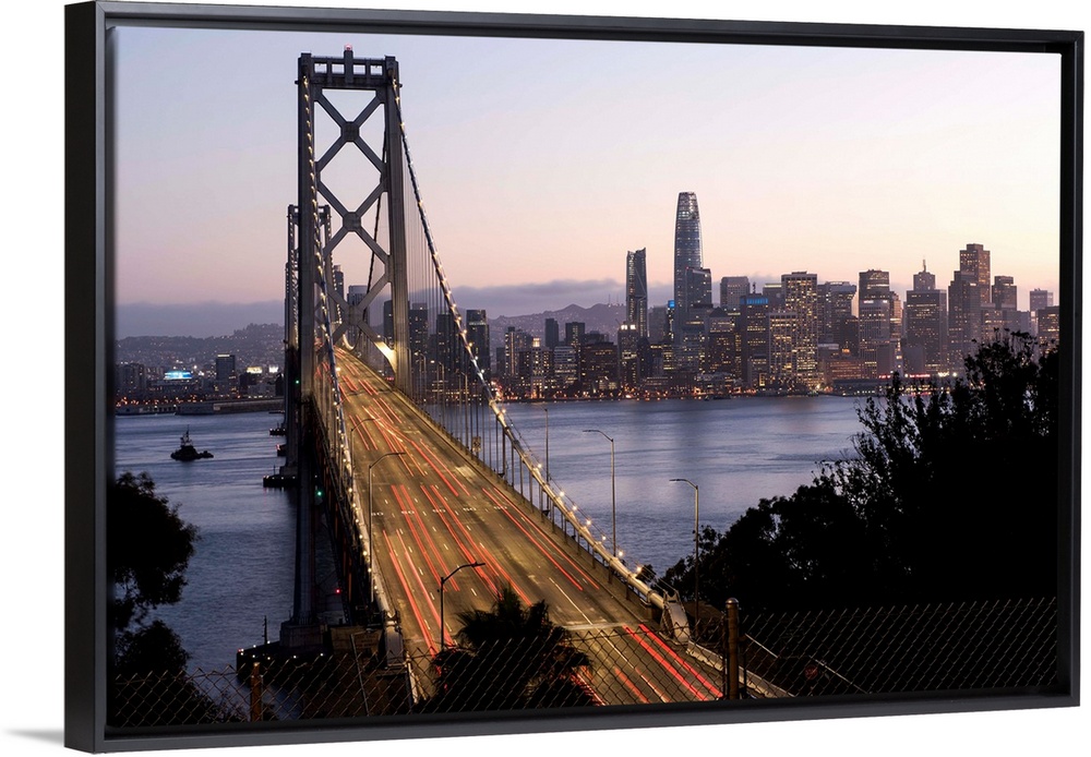 Photograph of the Bay Bridge with a pink and purple sunset and the San Francisco skyline lit up in the background.