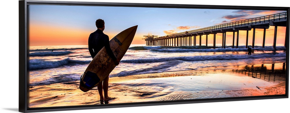 Silhouetted panoramic photograph of a man holding a surf board on the shore of Scripps Beach in San Diego, California, wit...