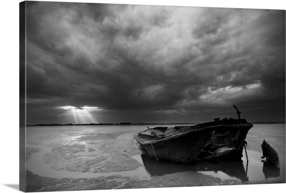 An abandoned, decaying boat sits in the sand under an overcast sky, with beams of light shining through in the distance.
