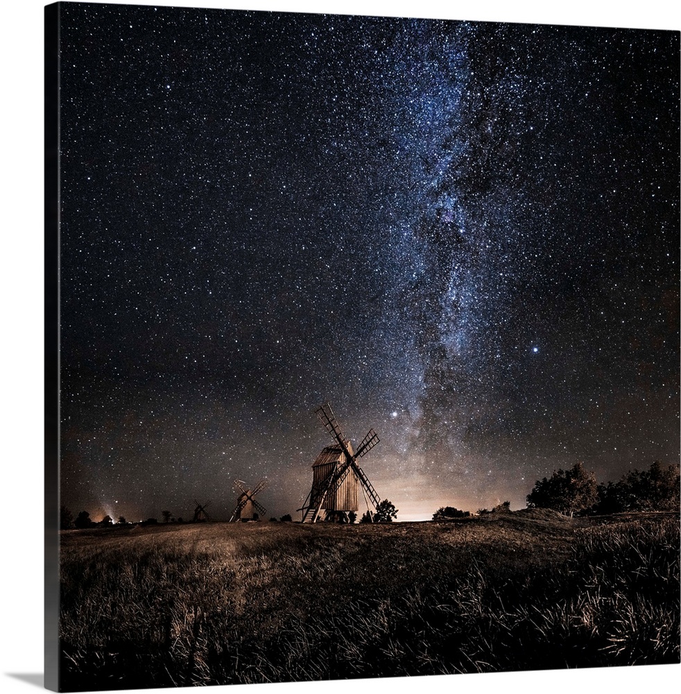 A dramatic photograph of a countryside scene with windmills in the distance and a starry night sky above.