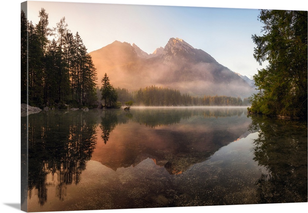 Reflective landscape photograph on a lake with trees and mountains in the background at sunrise.