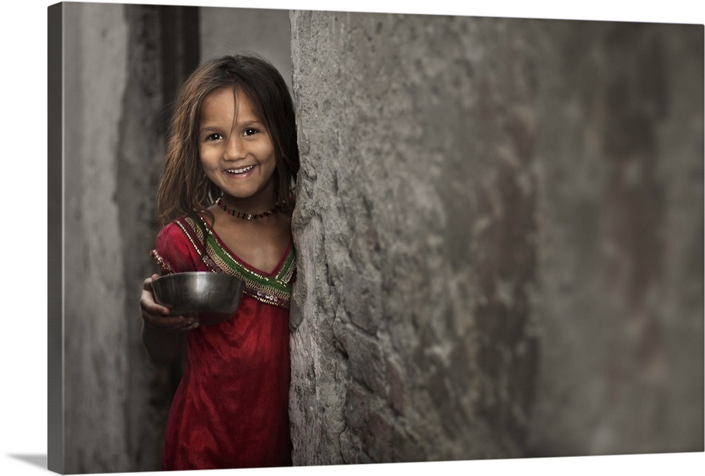 A smiling little girl holding a bowl, standing beside a stone wall.