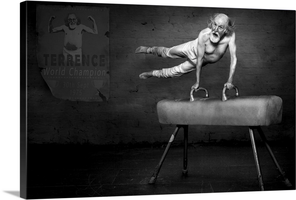 Black and white portrait of an elderly man on a pommel horse, with a poster showing his accomplishments on the wall behind...