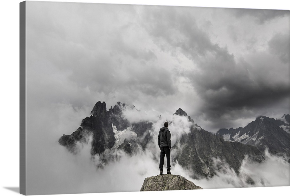 A portrait of a man standing on a large rock formation staring off into the distance at a cloudy mountain range.