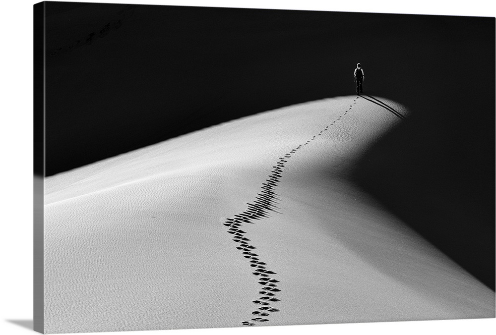 A man walking along the top of a sand dune into darkness leaving footprints behind him.