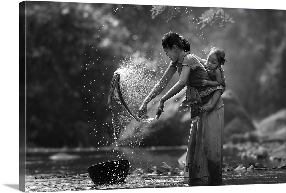 A mother in Indonesia with her child on her back washes clothing in a river.