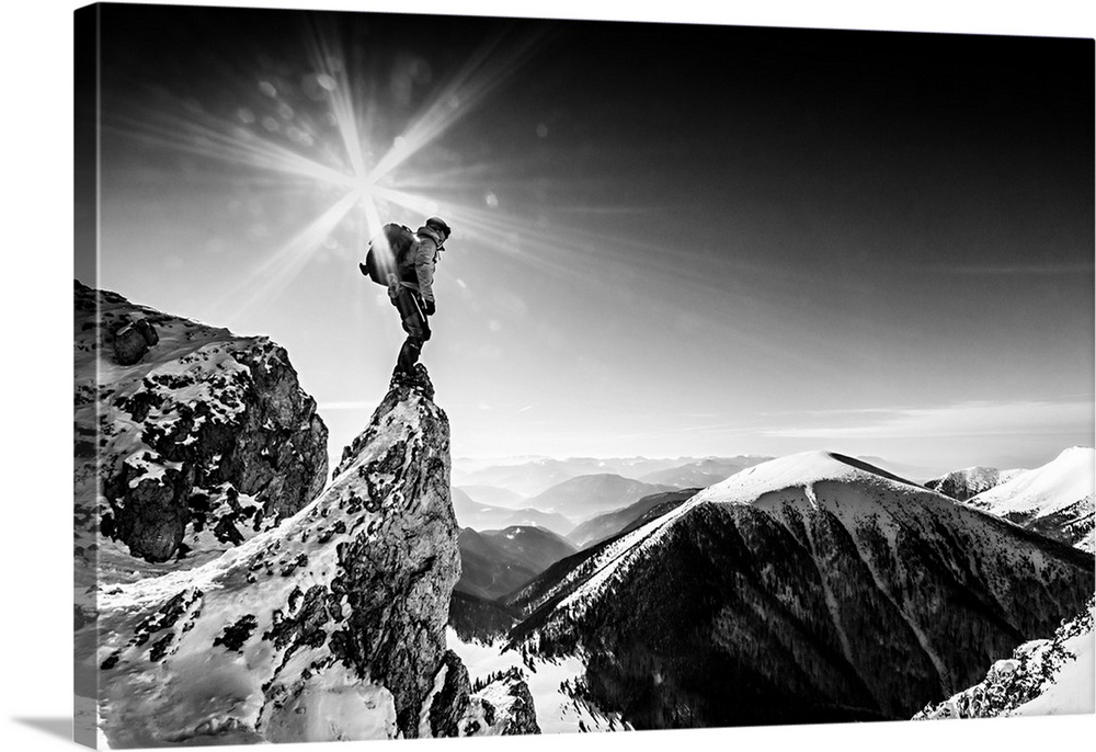 A sunburst shining over a hiker perched on the tip of a crag, overlooking a mountain valley.