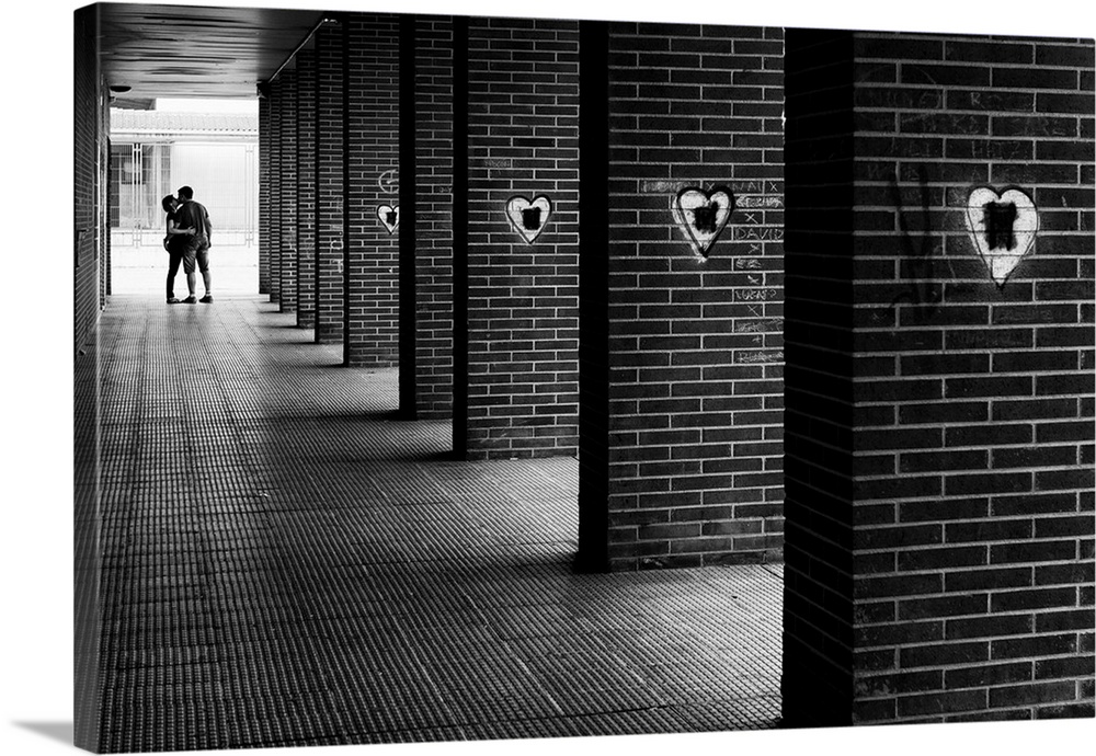 Black and white photograph with leading lines from brick columns with spray painted hearts on them and a couple kissing in...