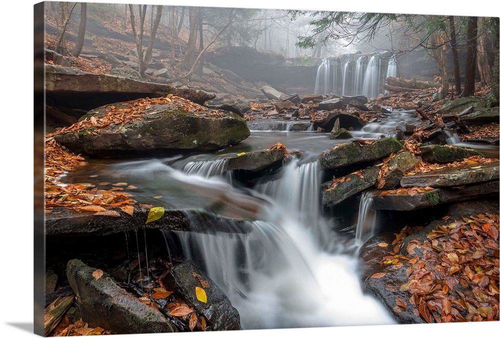 A waterfall and stream with a rocky bed and fallen leaves in a misty forest in Ricketts Glen, Pennsylvania.