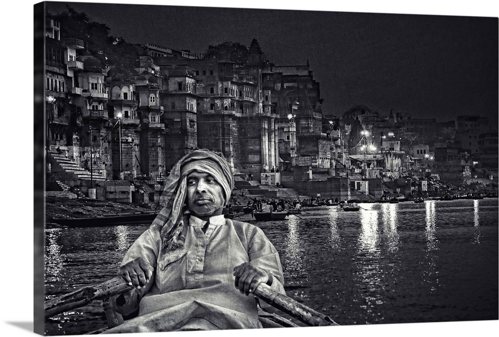 A man rows a boat on the river near an urban cityscape at night, India.