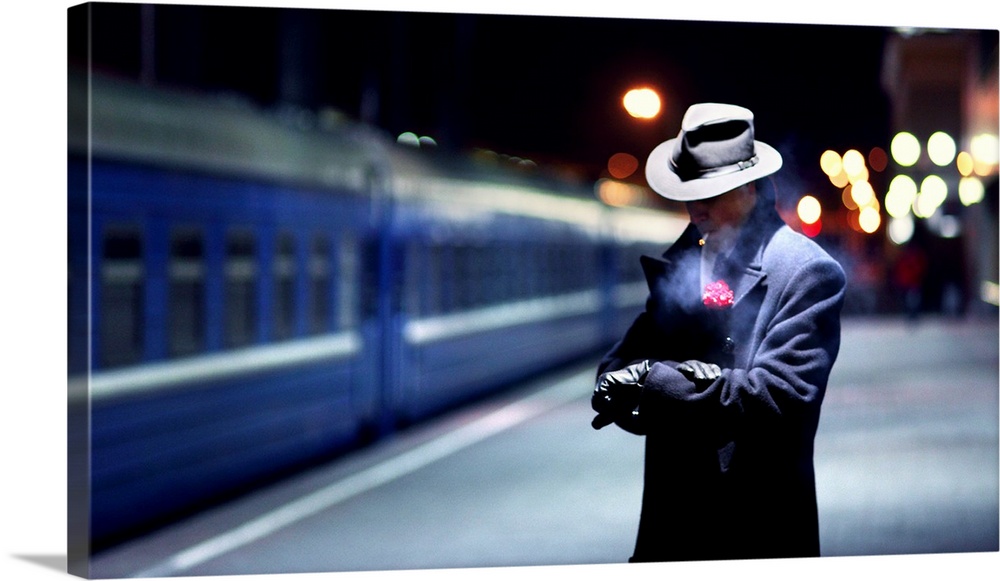 A man smoking a cigarette and wearing a boutonniere looking at his watch as a train passes by.