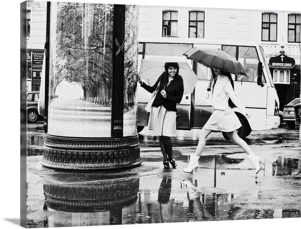 Two young women with umbrellas avoid puddles in the street.