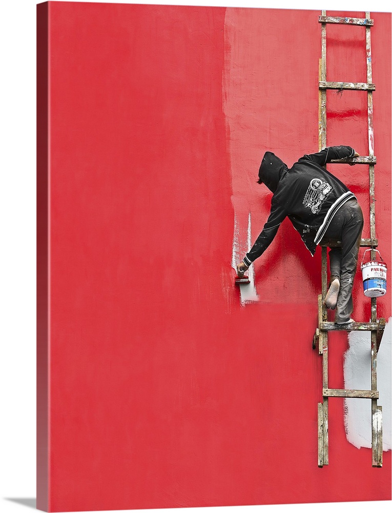 A person hanging from a ladder, painting the side of a building bright red.