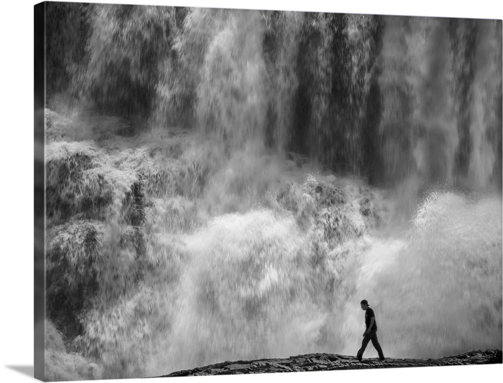 Man walking at the base of a huge water fall, looking tiny in comparison.