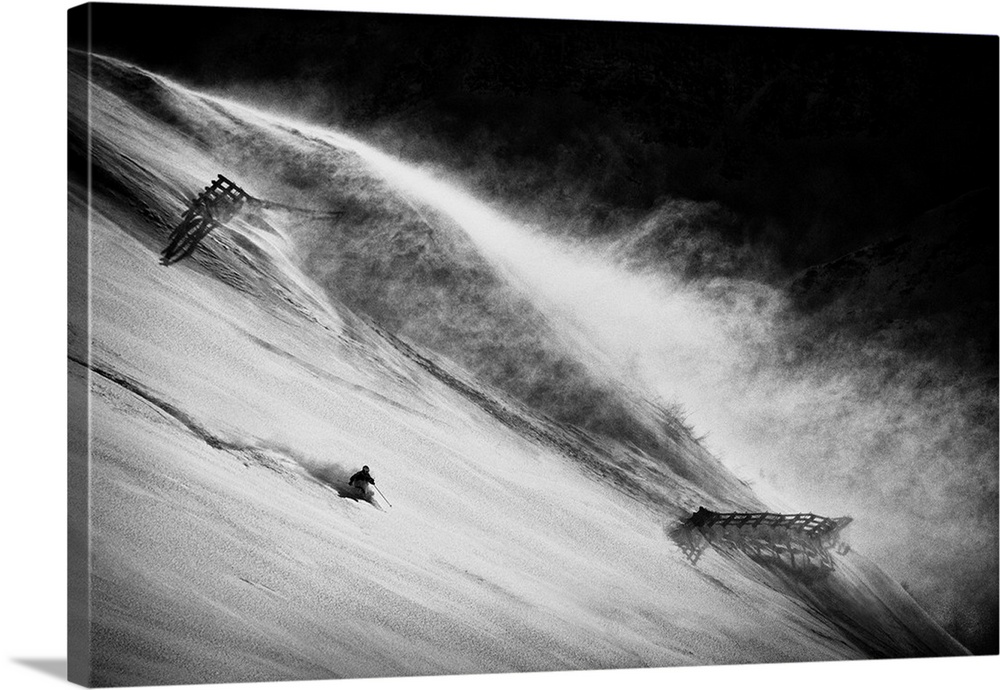 A skier heads down a slope past wooden fences, with wind blowing snow into the air.