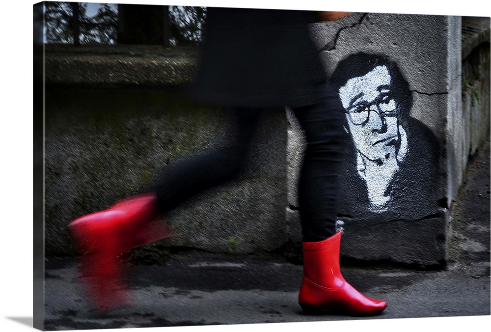 A woman in red boots walks quickly down a street past a stenciled image of Woody Allen on a concrete wall.