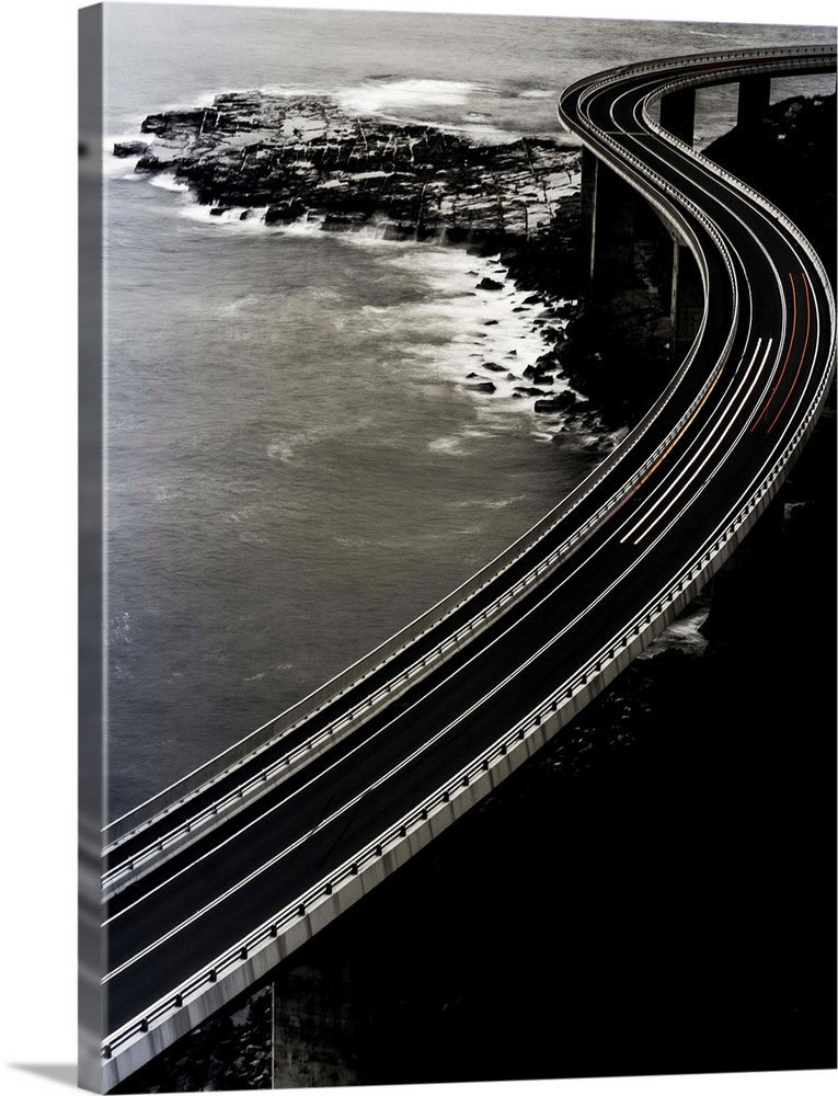 Vertical long exposure photograph of a winding bridge over an ocean with red and orange lines from traveling cars.