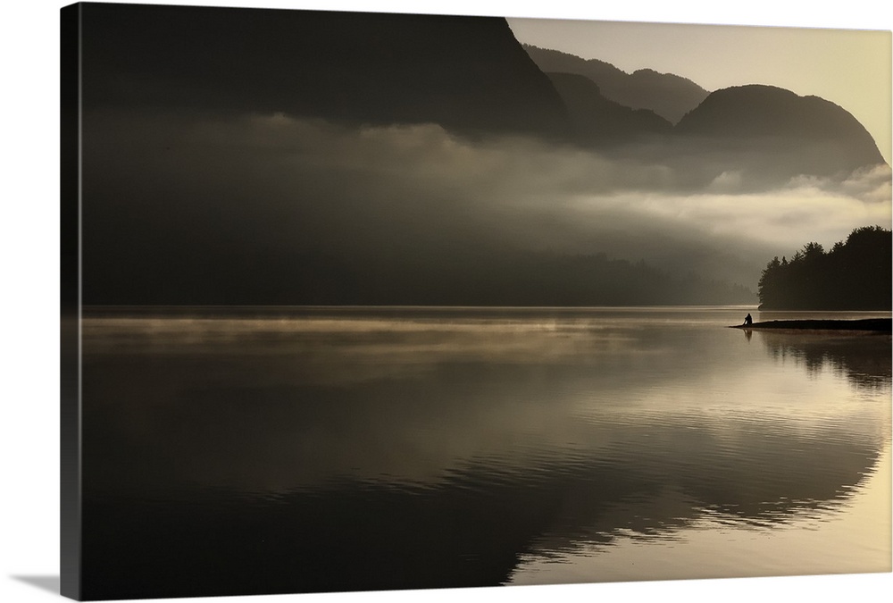 Distant figure sitting on the banks of a lake with misty clouds at sunset.