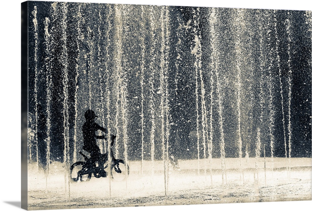A child pedals his bicycle through numerous water jets of a fountain.