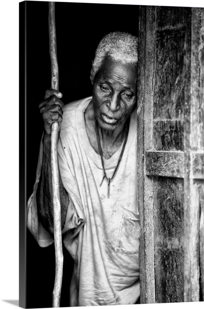 Black and white portrait of an elderly person with a staff in a doorway.