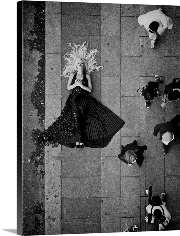A beautiful blond woman in a flowing dress laying on the sidewalk as people walk by.