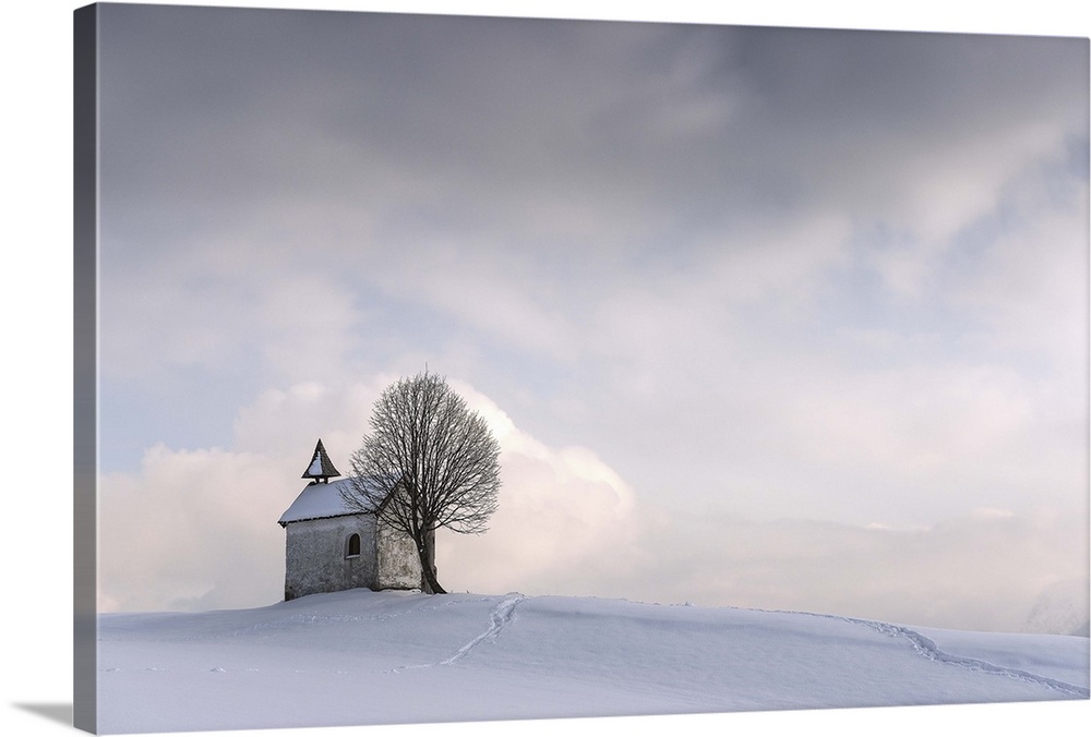 A small chapel with a tree sit on a hilltop covered in fresh snow in winter.