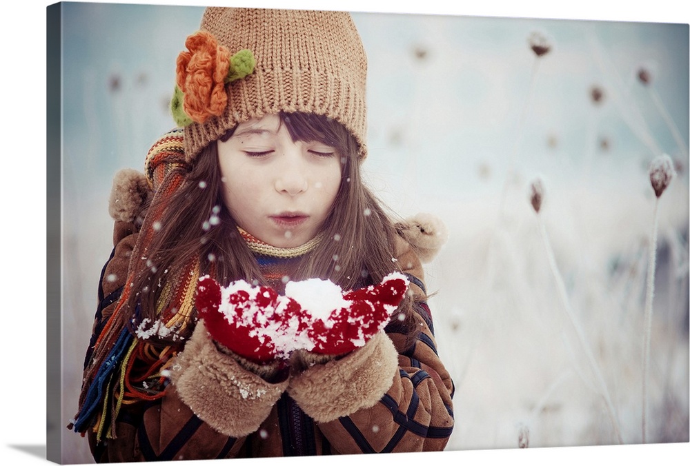 A young girl in winter clothes blows on a handful of snow.