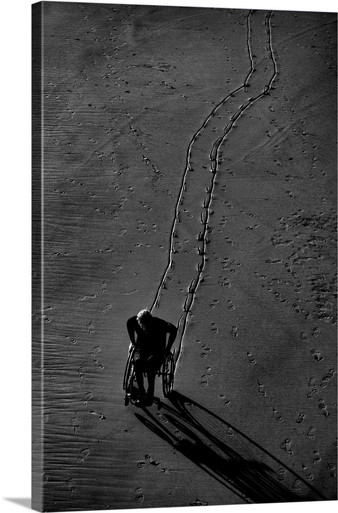Black and white photograph of a person using their wheel chair in the sand, leaving a lined trail amongst the footprints.