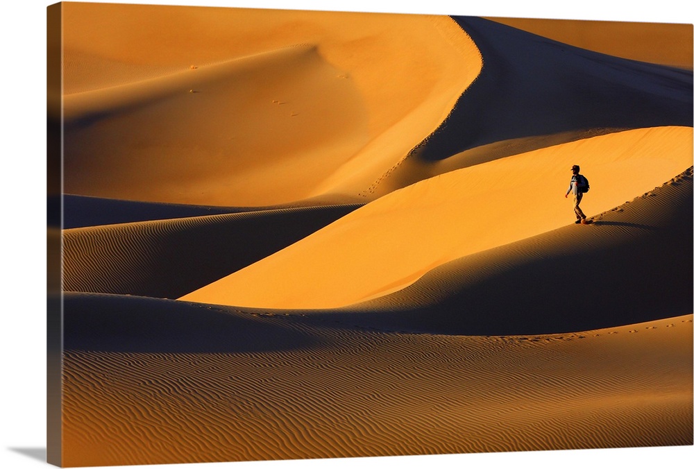 A person walks on a sand dune in a golden desert at sunset.