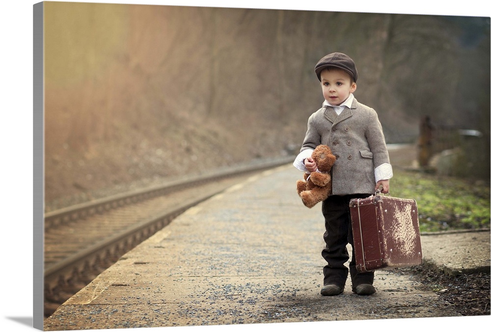 A young boy holding a teddy bear and a suitcase waits near train tracks.