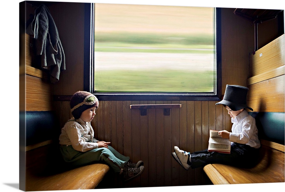 Two young boys, one in a dress shirt and wool hat, and the other in playclothes, sit opposite one another on a moving train.