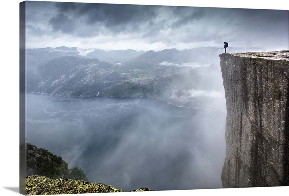 A hiker stands at the edge of a steep cliff overlooking a misty fjord, Prekestolen, Norway.