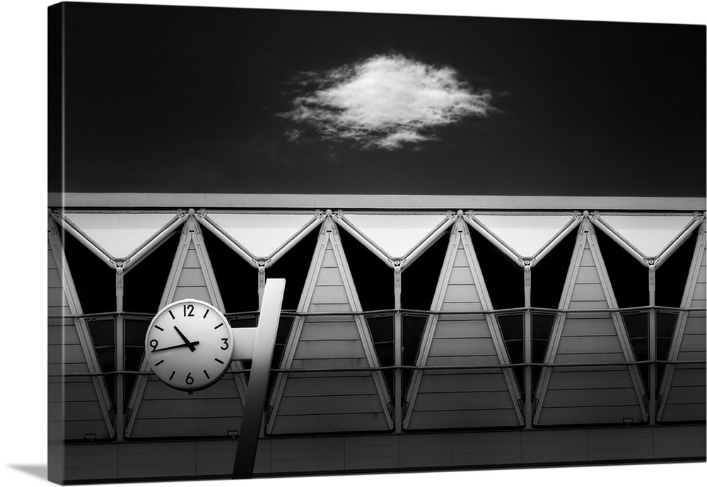 Black and white abstract photograph of a clock hung in front of unique triangular windows with a single cloud in the sky a...