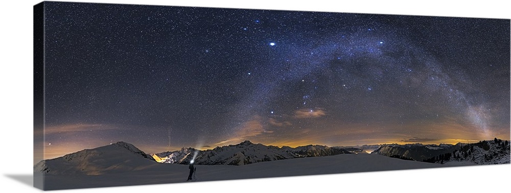 A panoramic photograph of a person standing in a desolate mountainous snowscape under a starry night sky, with the milky w...