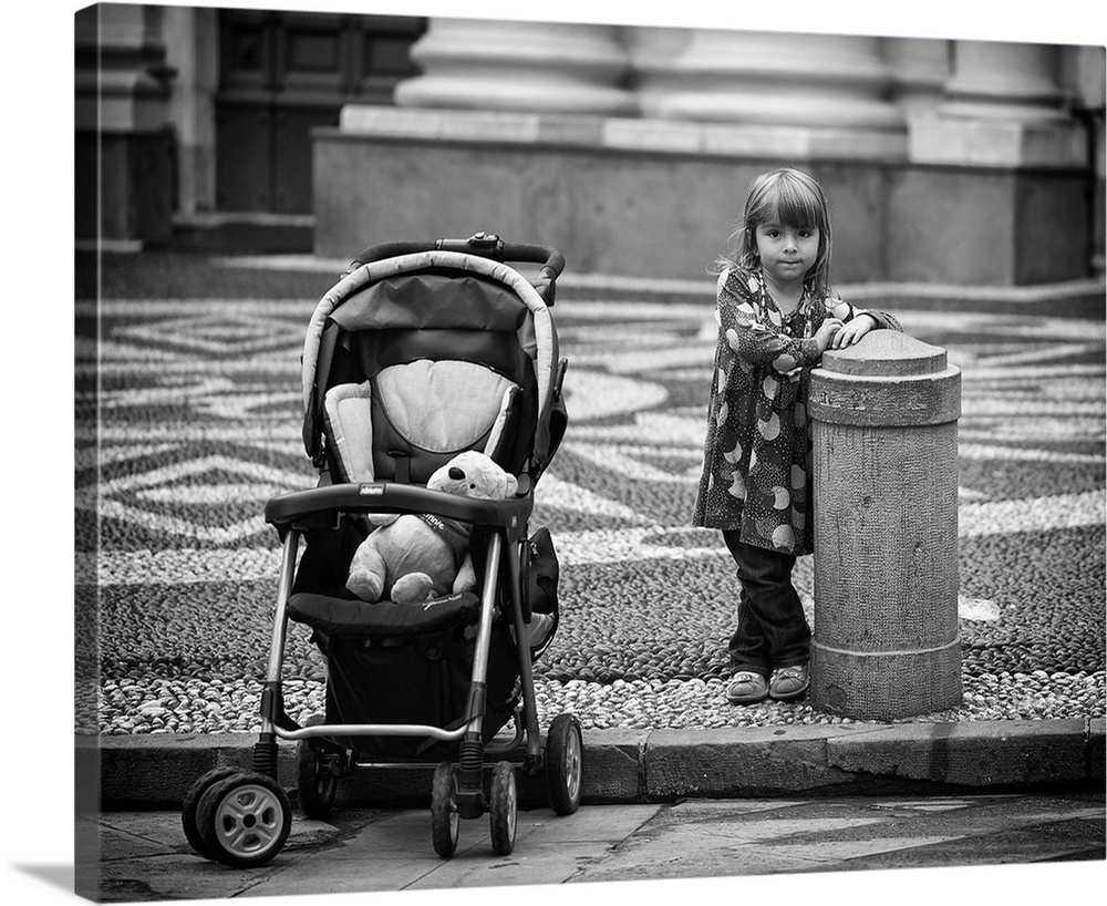 A little girl waits next to a cement barrier on the street with her teddy bear in her stroller.