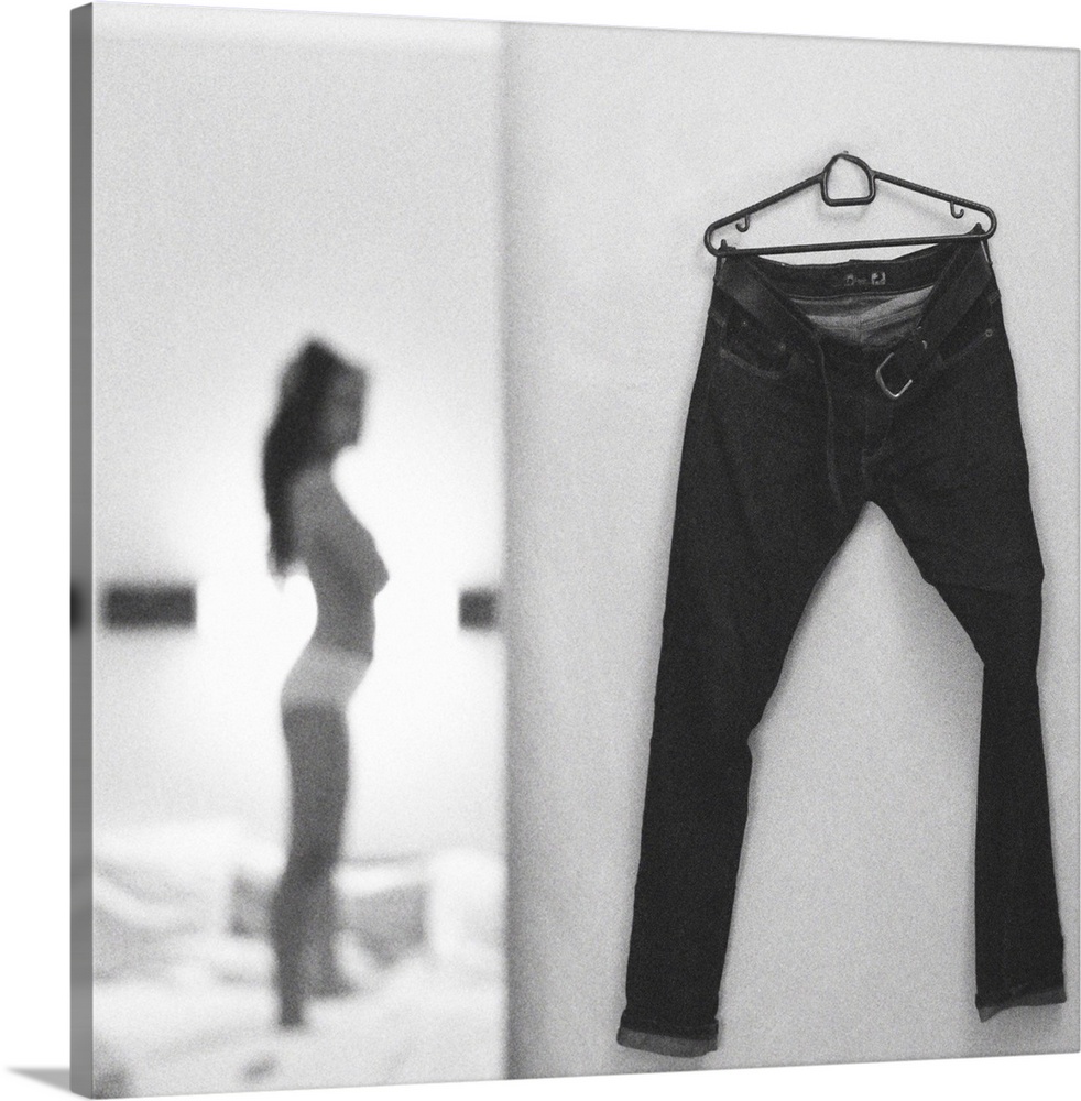 A focus on a pair of pants hanging on a wall while a young woman in the background gets ready to put them on.