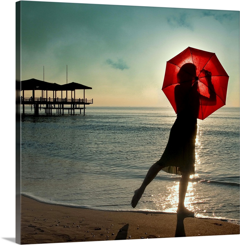 A woman on the beach seen through a red umbrella, with a pier in the distance.