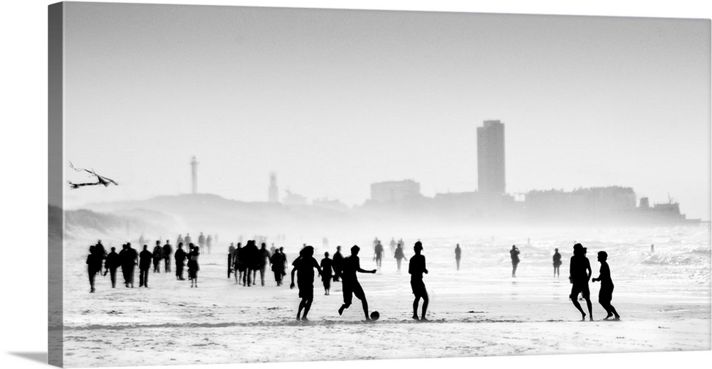 Soft, horizontal black and white photograph with silhouettes of people walking and playing on the beach with a skyline in ...
