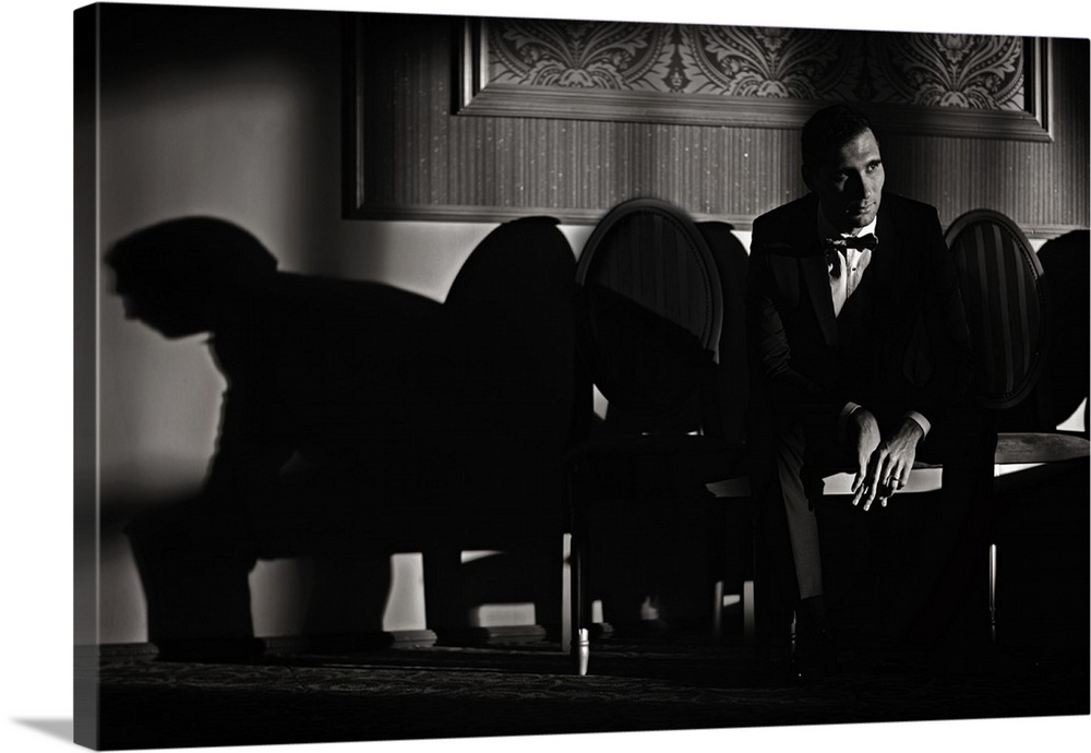 A man sits in a chair in a tuxedo casting long shadows on the wall.
