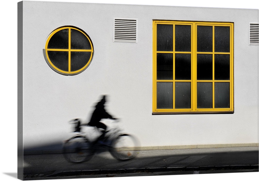 A person rides by a white wall with yellow trim windows on bicycle.