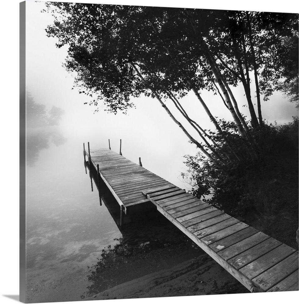Black and white photograph of a dock jetting out over a foggy lake in the countryside.