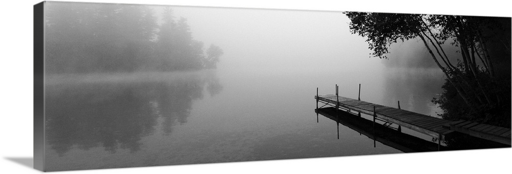 A black and white photograph of a dock stretching out over a foggy lake in an idyllic setting.