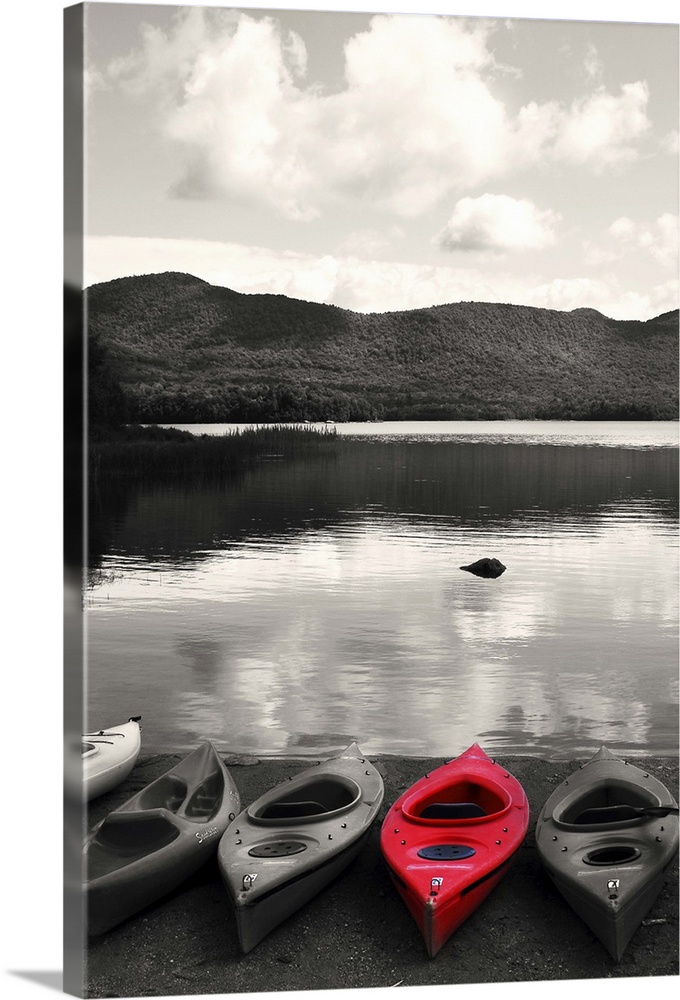 Black and white photograph with a bright red kayak next to other kayaks, resting on the shore of a lake.