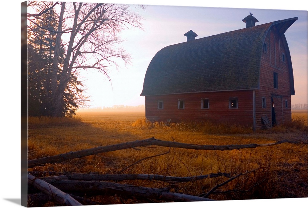 A large barn is photographed in the morning during sunrise and there is a thin layer of fog over the land.