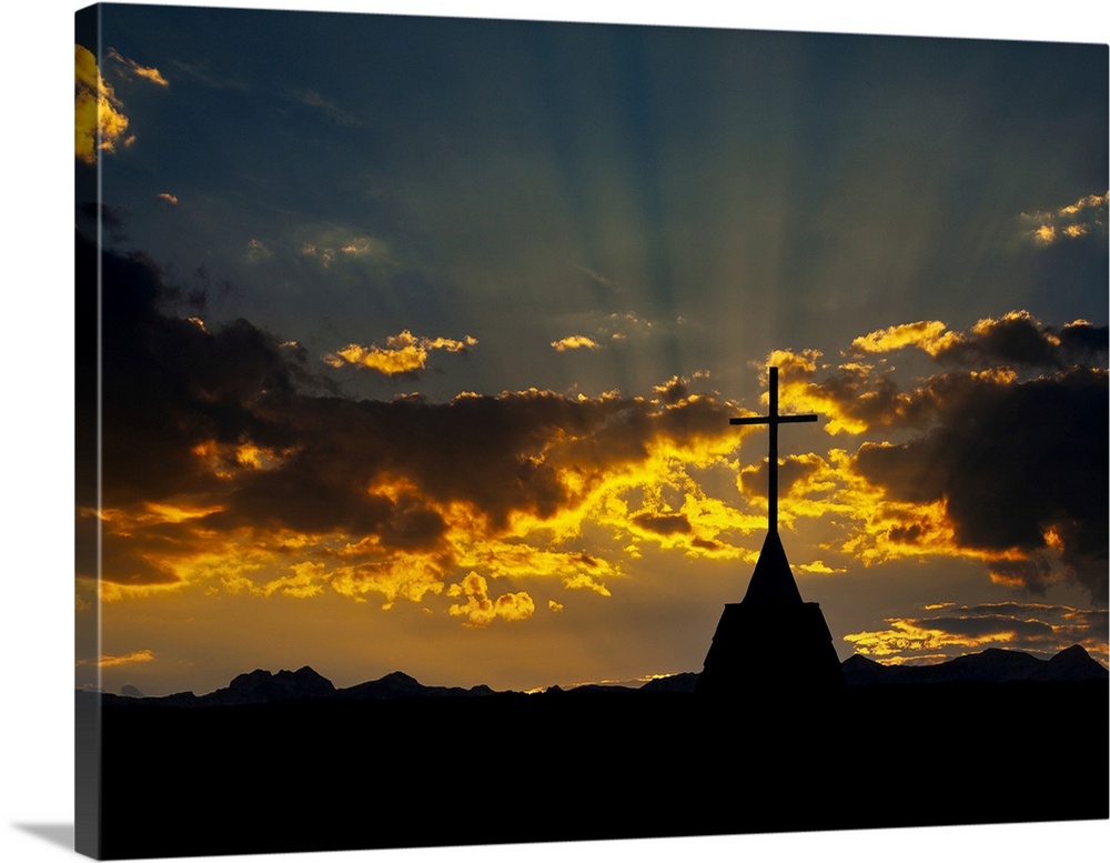 Silhouette of a church steeple's cross against a dramatic colorful sky with silhouette mountain range in the background. C...