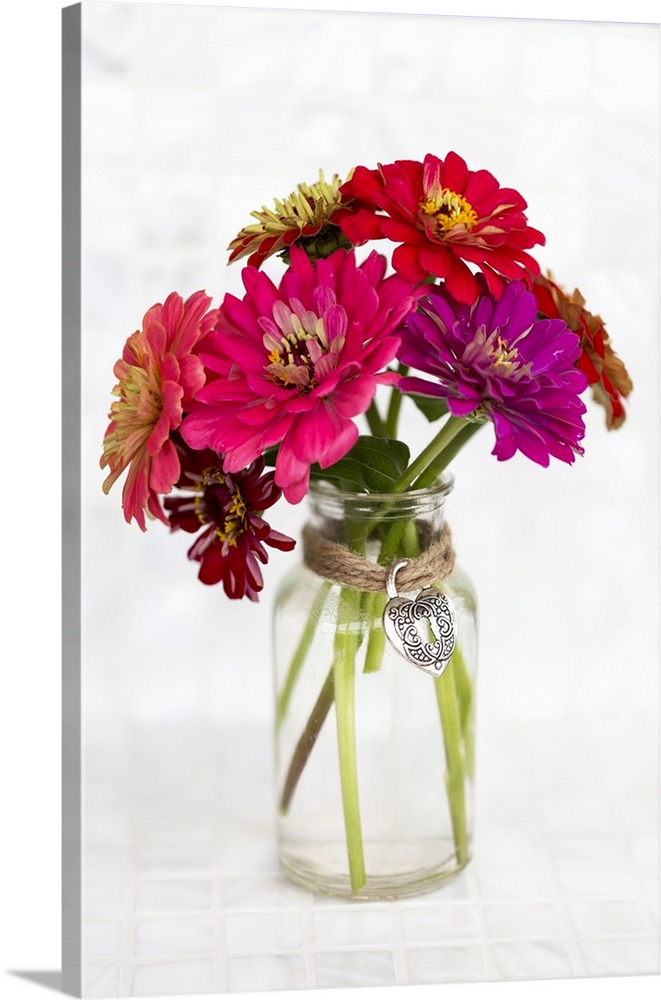 A variety of colored Zinnia flowers in a simple glass vase with a decorative heart pendant held in place with a jute string