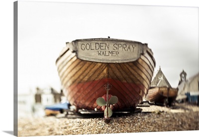 A wooden motorboat sits on the shingle beach with buildings in the background; England