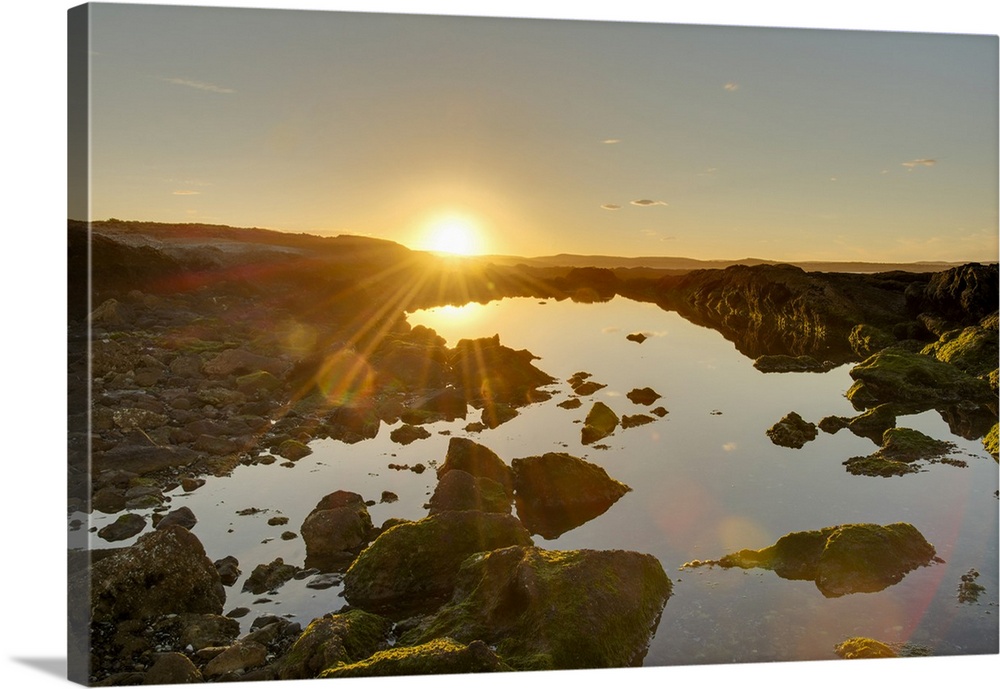 A golden-coloured sunset and sunburst hilights the rocks around an ocean-side pool of water; Chubut, Argentina