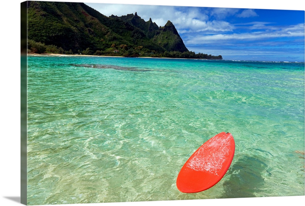 Photograph of bobbing surfboard in ocean with crystal clear waters and a small beach with tree covered mountains in the di...