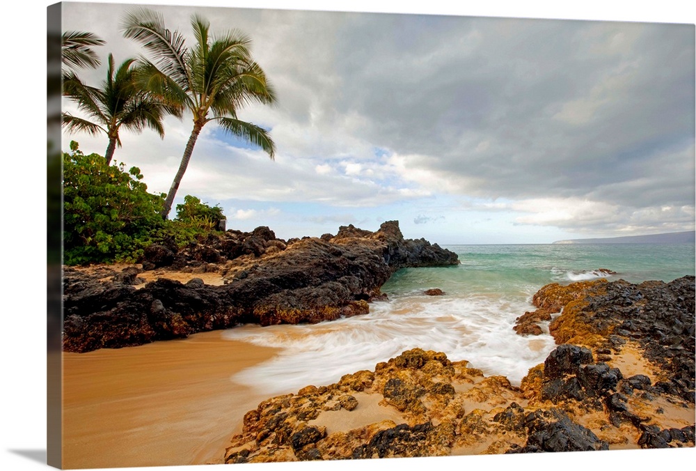 Photograph of a sandy break in the rocky coast as water rushes in to the cove with palm trees on an overcast day.
